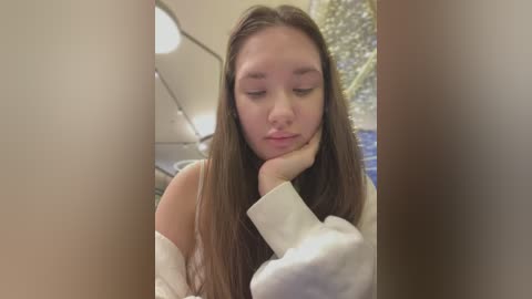 Media: A video of a young woman with long brown hair, wearing a white sweater, resting her head on her hand, indoors with blurred background.