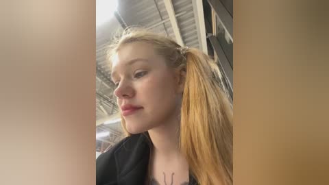 Media: Video of a young blonde woman with long hair tied in a high ponytail, wearing a black jacket, standing in an industrial building with a high ceiling and metal beams.