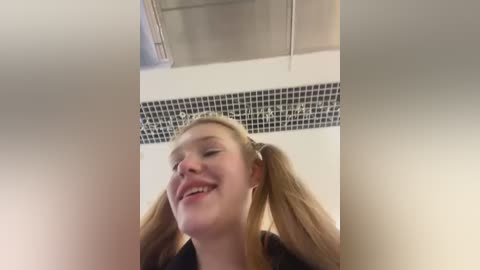 Media: A video of a smiling, young, fair-skinned woman with blonde pigtails, wearing a black top, standing in a modern bathroom with a white tiled wall and black mosaic border.