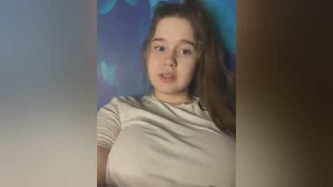 Video of a young, fair-skinned woman with long brown hair, wearing a beige t-shirt, set against a blue and purple gradient background. She has a soft expression, possibly mouthing something.