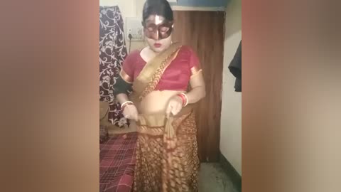 Media: A video of a woman dressed in traditional Indian attire, including a red blouse, gold sari, and a black mask, standing in a dimly lit room with patterned curtains and a wooden door in the background.