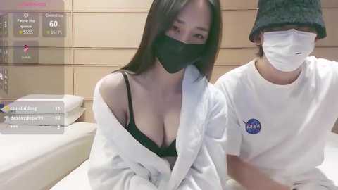 Media: Video of two Asian women, one in a black bra, wearing a face mask, sitting on a bed, the other in a white shirt and face mask, in a hospital room.