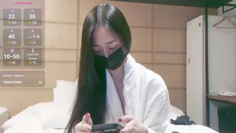 Media: A video of an East Asian woman in a white robe, wearing a black mask, reading a phone, with a temperature display and white bed in the background.