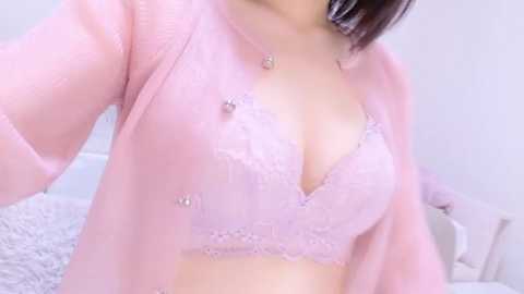 Video of an East Asian woman with light skin, wearing a pastel pink lace bra and matching cardigan, with a hint of her breasts visible. The background shows a white bed and a fluffy white blanket.