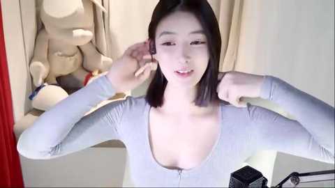 Media: Video of a young Asian woman with straight, shoulder-length black hair, wearing a light grey, long-sleeve top with a deep neckline, adjusting her earpiece in a modern, minimalist room with beige curtains and plush toys.