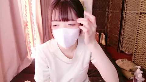 Media: Video of an Asian woman with straight black hair, wearing a white mask, adjusting her glasses. She's in a cozy room with beige curtains, wicker screen, and a dark sofa.