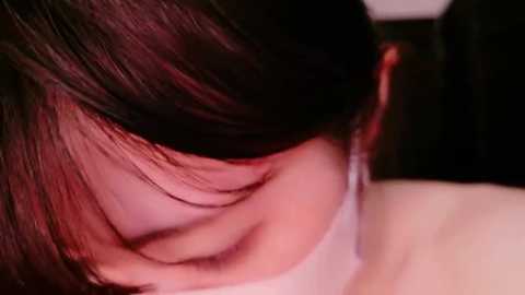 Media: A close-up video of a person with shoulder-length, dark brown hair and fair skin, wearing a white choker necklace. The image is dimly lit, highlighting the hair and skin texture.