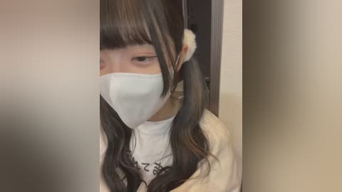 Media: A video of an East Asian woman with long, dark hair, wearing a white mask, beige sweater, and a light-colored jacket, partially obscured by a frosted glass door.