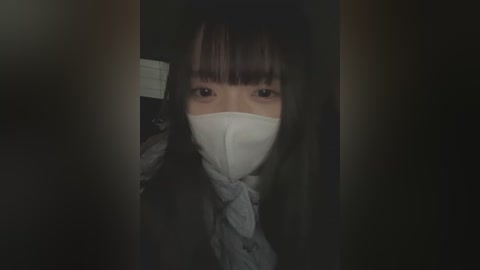 Media: Video of an Asian woman with long, straight black hair, wearing a white surgical mask and a gray coat, standing in a dimly lit, dark room.