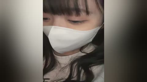 Media: Video of a young Asian woman with long, straight black hair, wearing a white surgical mask, and a beige sweater, blurred background.