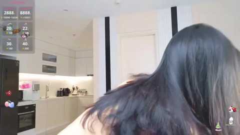 Media: Video of a person with long black hair, taken from behind in a modern kitchen with white cabinets, a black fridge, and a counter with appliances.