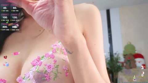 Media: Video of a light-skinned woman in a pink floral bra, with her arm raised, revealing her cleavage. Background shows a cluttered room with a potted plant and a green lamp.