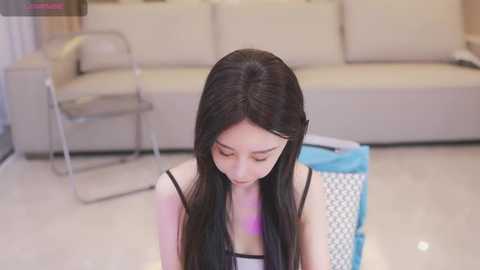 Media: A video of a young Asian woman with long black hair, wearing a black spaghetti-strap top, kneeling on a glossy white floor in a modern living room with beige furniture and a blue basket.