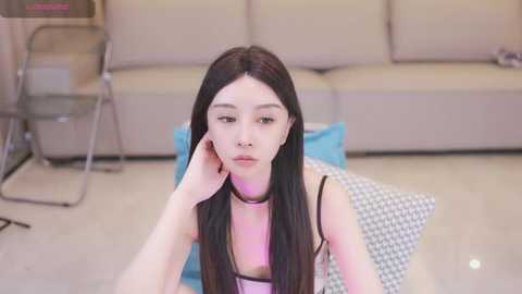 Media: Video of an Asian woman with long black hair, wearing a black choker and a pink tank top, sitting on a houndstooth cushion in a modern, beige-lit living room.