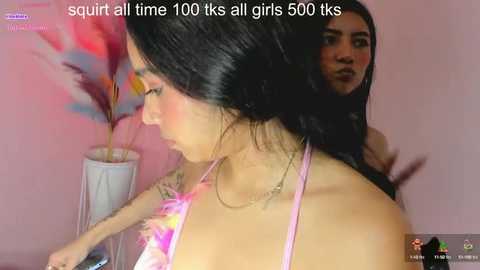 Media: A video of a young woman with long black hair, wearing a colorful tie-dye top, and a necklace, standing against a pink wall. A second woman with long black hair is seen in the background.