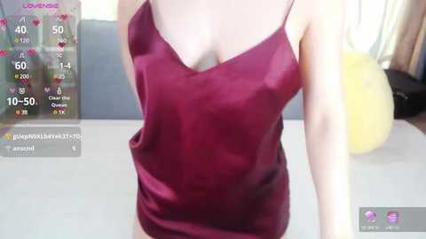 Media: Video of a woman in a maroon satin camisole with thin straps, showcasing a small bust, against a light-colored background. The image includes a smartphone screen overlay displaying user interface elements.