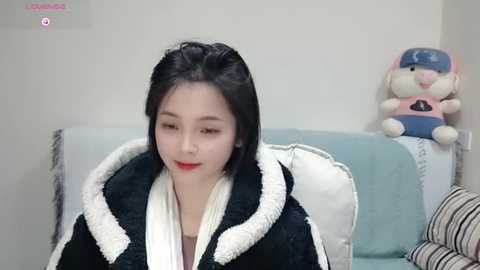 Media: Video of a young East Asian woman with fair skin and shoulder-length black hair, wearing a black and white fur-trimmed robe, sitting on a striped couch in a minimalist room. A plush toy resembling a cat sits on a nearby cushion.