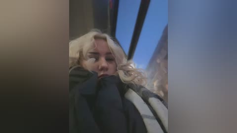 Media: Video of a blonde woman, possibly in her 20s, wearing a black coat, sitting in a car with a blurred, dark window.