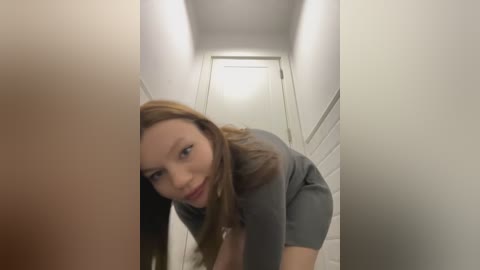 Media: Video of a young Caucasian woman with long brown hair, wearing a gray dress, leaning forward, looking directly at the camera, in a narrow, dimly lit hallway with white walls and a closed door.