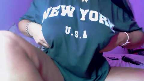 Media: Video of a woman wearing a dark green \"NEW YORK U.S.A.\" t-shirt, lifted to reveal her black underwear. She has a light skin tone and a thin build.