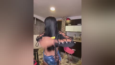 Media: Video of a woman with long black hair, wearing a black lace top and denim shorts, in a modern kitchen with a stone wall, wooden cabinets, and a patterned rug.