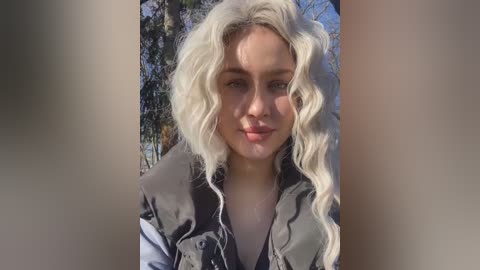 Media: Video of a fair-skinned woman with long, wavy platinum blonde hair, wearing a dark leather jacket, standing outdoors amidst tall trees and sunlight.