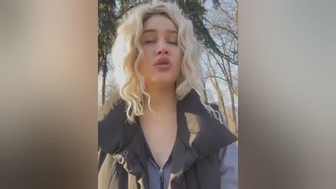 Media: Video of a young, fair-skinned woman with blonde, wavy hair, wearing a black puffer jacket, puckering her lips in a kiss, outdoors against a backdrop of tall, bare trees in sunlight.