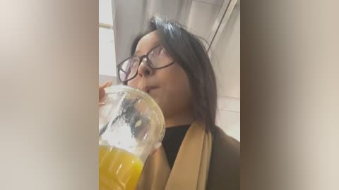 Media: Video of a woman with shoulder-length black hair, wearing glasses and a beige coat, drinking from a plastic cup filled with orange juice. She is indoors, near a window with white curtains.