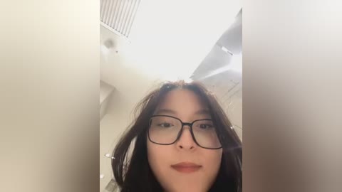 Media: A video of an Asian woman with long dark hair, wearing black-rimmed glasses, standing inside a small, white, narrow space with a vented ceiling and beige walls.