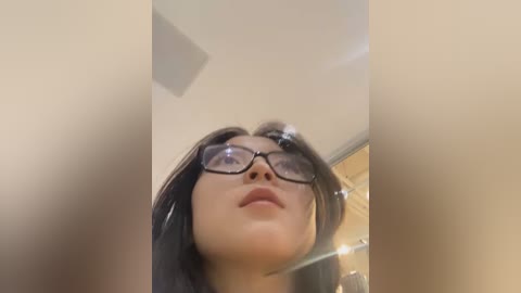 Media: Video of a young woman with medium skin tone, long black hair, and large black glasses, looking upward in a dimly lit room with beige walls and a ceiling light.