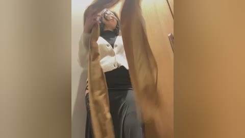 Media: Video of a woman with medium-brown skin and shoulder-length dark hair, wearing a white cardigan and black skirt, holding a beige purse, standing in a narrow hallway with beige walls.