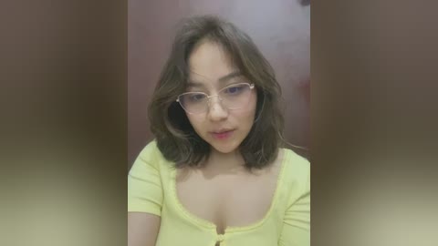 Media: Video of an Asian woman with shoulder-length brown hair, wearing glasses and a yellow blouse with a deep neckline, against a blurred brown background.