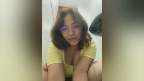 Media: Video of an Asian woman with shoulder-length brown hair, wearing glasses and a yellow top, sitting on a tiled floor, smiling.