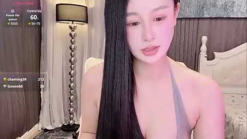 Media: A video of an East Asian woman with long black hair, wearing a halter top, standing in a bedroom with a white bed, lamp, and gray walls.