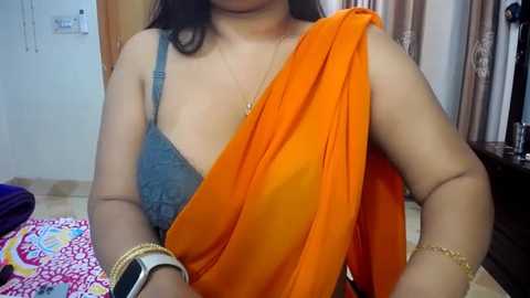 Media: Video of a woman with medium skin tone wearing a blue lace bra and orange saree, in a home setting with a white wall, wooden door, and a pink patterned bedspread.