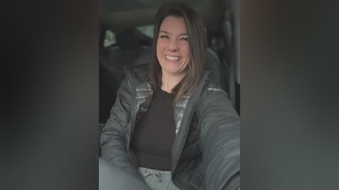 Media: Video of a smiling, young Caucasian woman with shoulder-length brown hair, wearing a black top and gray puffer jacket, sitting in a car's back seat.