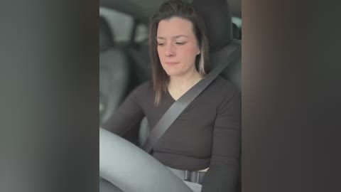 Media: Video of a woman with light skin, straight brown hair, wearing a black long-sleeve shirt, seated in a car with a gray seatbelt, looking down with a neutral expression.