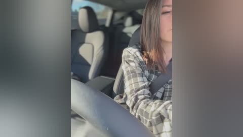 Video of a woman in a car, wearing a plaid shirt, eyes closed, seated in the driver's seat, with blurry surroundings.