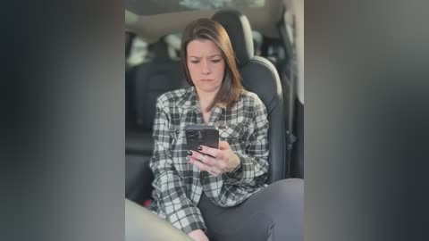 Media: Video of a woman with shoulder-length brown hair, wearing a plaid shirt, sitting in a car, intensely focused on her smartphone.