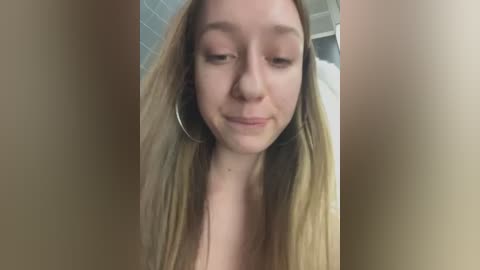 Media: Video of a young Caucasian woman with light skin, long blonde hair, and a slight smile. She wears large hoop earrings and is partially nude, with her breasts visible. The background is blurry, showing a tiled bathroom wall.