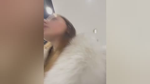 Media: A blurry video of a woman with long hair, wearing a white fur coat, leaning against a white wall.