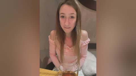 Media: A video of a young woman with long, straight brown hair, wearing a pink off-shoulder top, sitting at a table with a glass of alcohol, surrounded by blurred, warm-toned background.