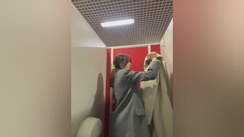 Video of a woman in a light gray coat hanging up a beige coat in a narrow, white-walled restroom with a red door and a grid-patterned ceiling light.