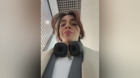 Media: Video of a young woman with light brown hair and light skin, wearing a beige jacket over a white shirt, holding black headphones, standing in a modern corridor with a grid-patterned wall.