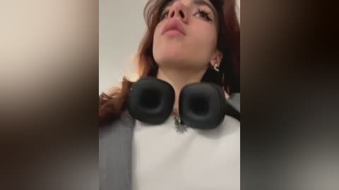 Media: Video of a young woman with light skin and long, wavy brown hair, wearing black over-ear headphones and a white sweater, looking upwards with a relaxed expression.