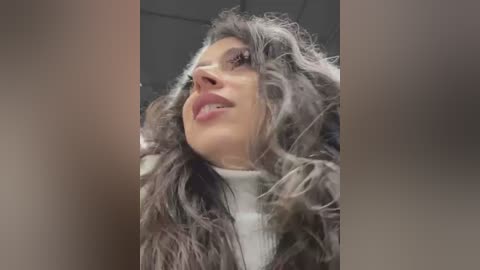 Media: Video of a close-up, side view of a woman with curly, voluminous gray hair and fair skin, wearing a white turtleneck sweater. Background is blurred, showing a ceiling with light fixtures.