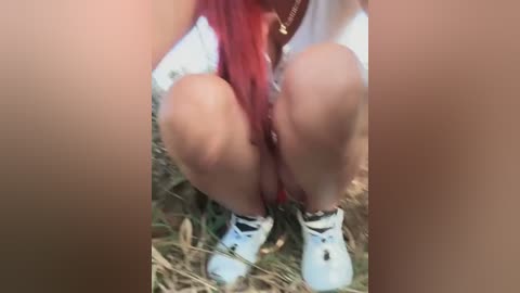 Media: A video of a person squatting outdoors with red hair, wearing white sneakers and shorts, holding a red object. The background is blurry, with grass and dirt.