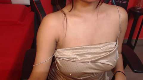 Media: Video of an Asian woman with fair skin, wearing a shimmering gold sequin dress with thin straps, sitting in a red cinema seat. Her dark hair is tied back.