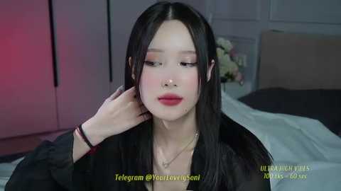 Media: A video of an East Asian woman with long black hair, red lipstick, and fair skin, wearing a black top, standing in a modern bedroom with white walls and a bed.