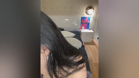 Media: Video of a woman with long black hair lying on a bed in a dimly lit bedroom. The room features a white headboard, a blue painting, and a wooden floor.